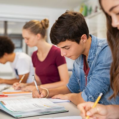 Group of college students studying in classroom writing notes during lesson. Focused guy and girls studying in college library sitting at desk. Group of multiethnic university students doing research sitting in a row.