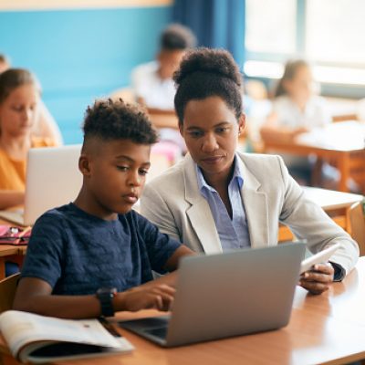Black female teacher assisting schoolboy in using laptop during a class in the classroom.